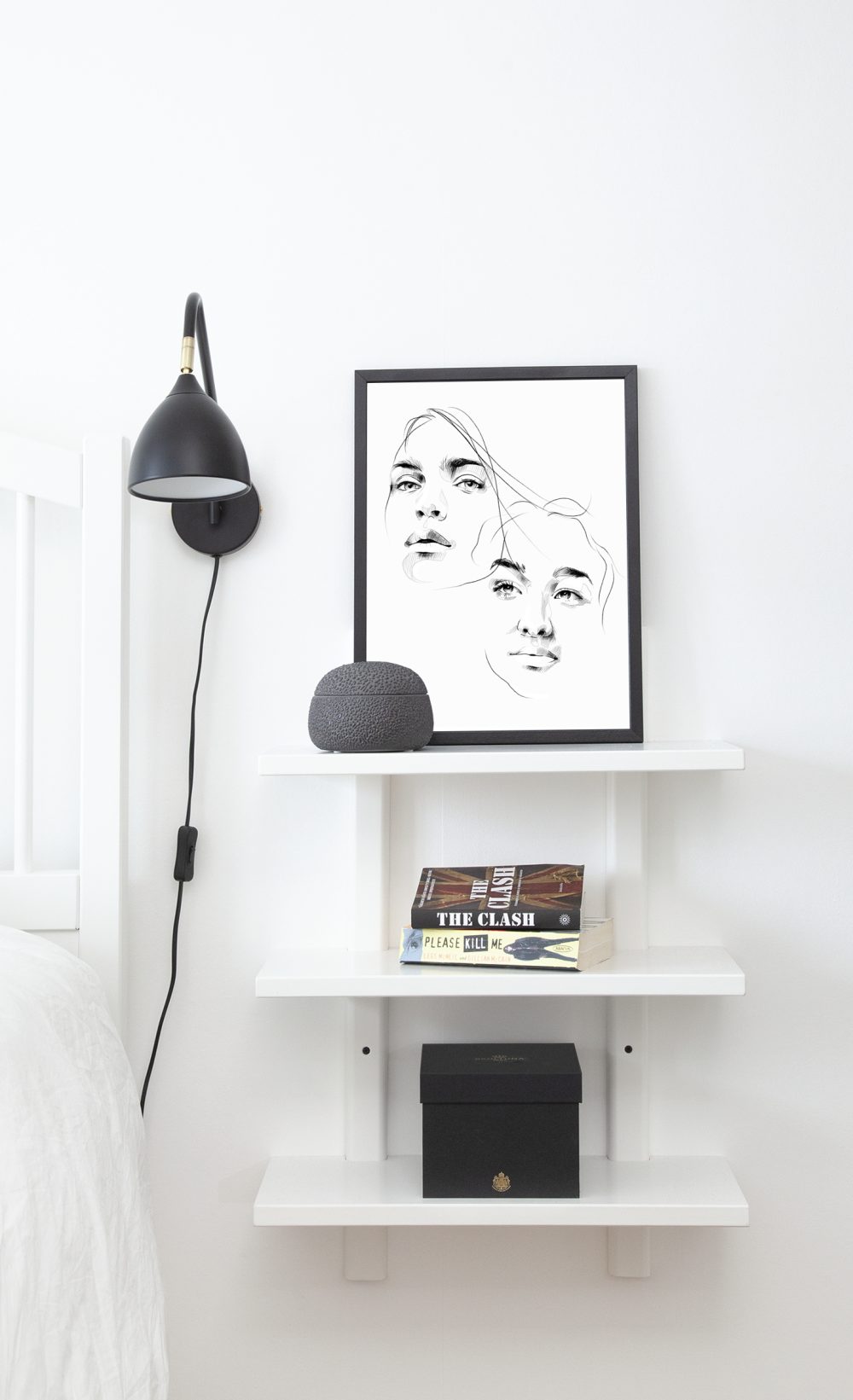 Art print with two portraits with black frame on a white shelf next to a lamp and bed.
